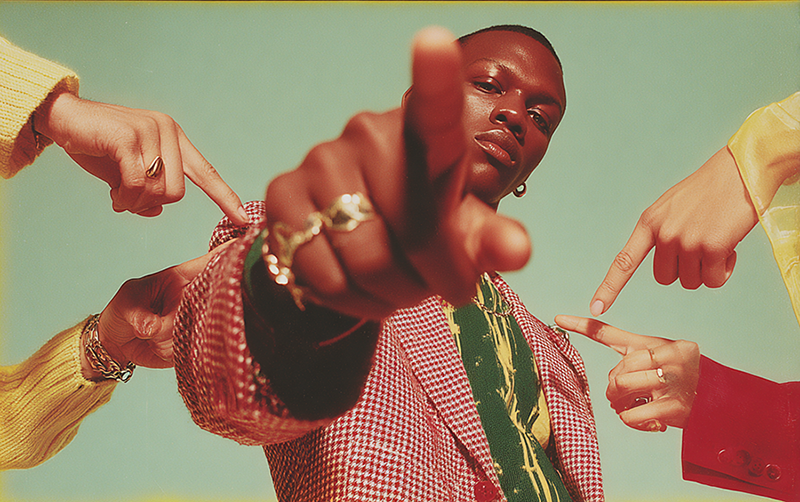 A man in a red check jacket stares directly ahead, pointing outward as others point at him — a visual metaphor for self-consciousness and the awareness of being seen.
