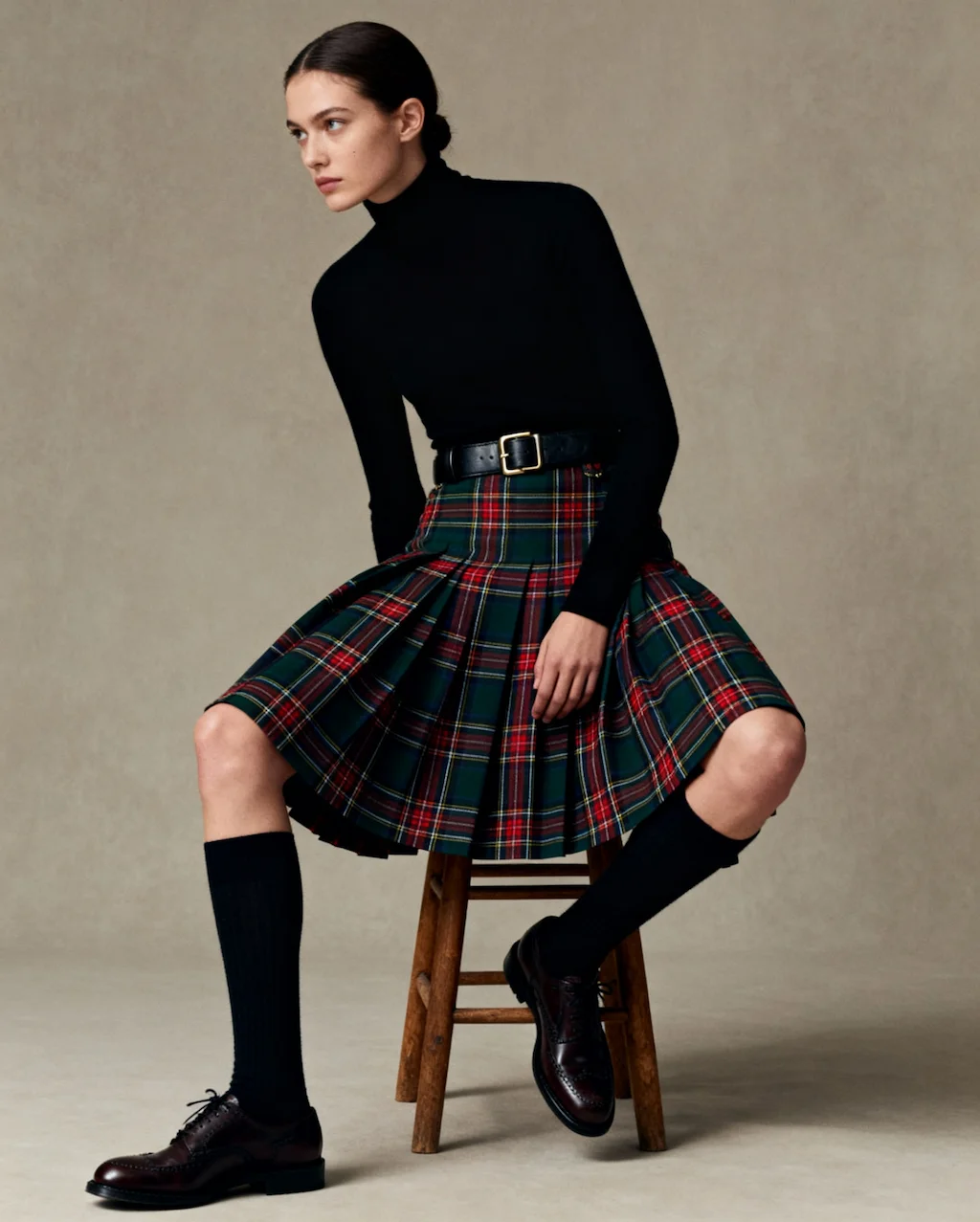A woman seated on a stool wearing a black turtleneck and a red-green tartan skirt in a classic daily outfit