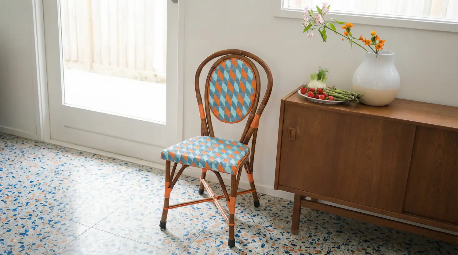 Scandinavian-style interior featuring a rattan chair with orange-blue woven pattern, a wooden cabinet, terrazzo flooring, and decorative flowers